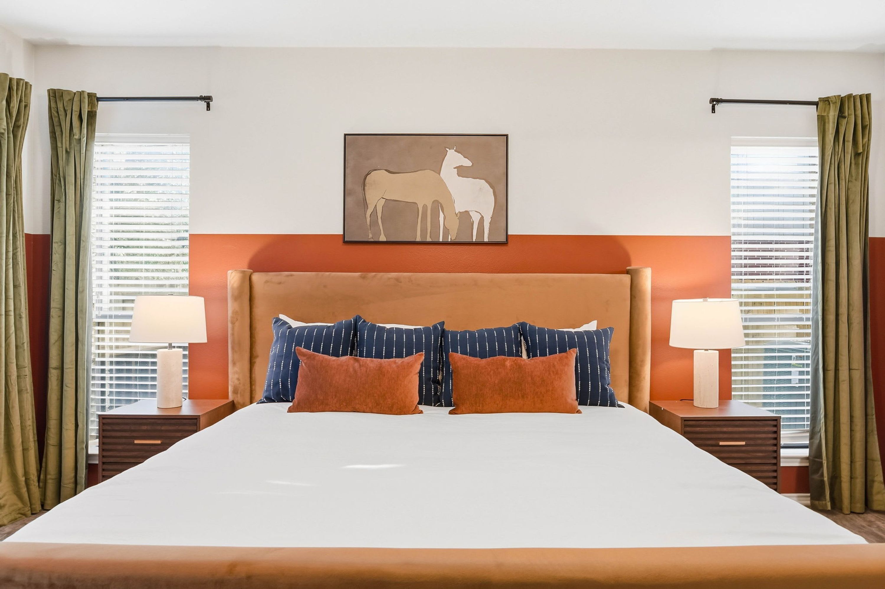 A hotel room with a large bed, two bedside tables with lamps, a headboard, and a painting of two animals.
