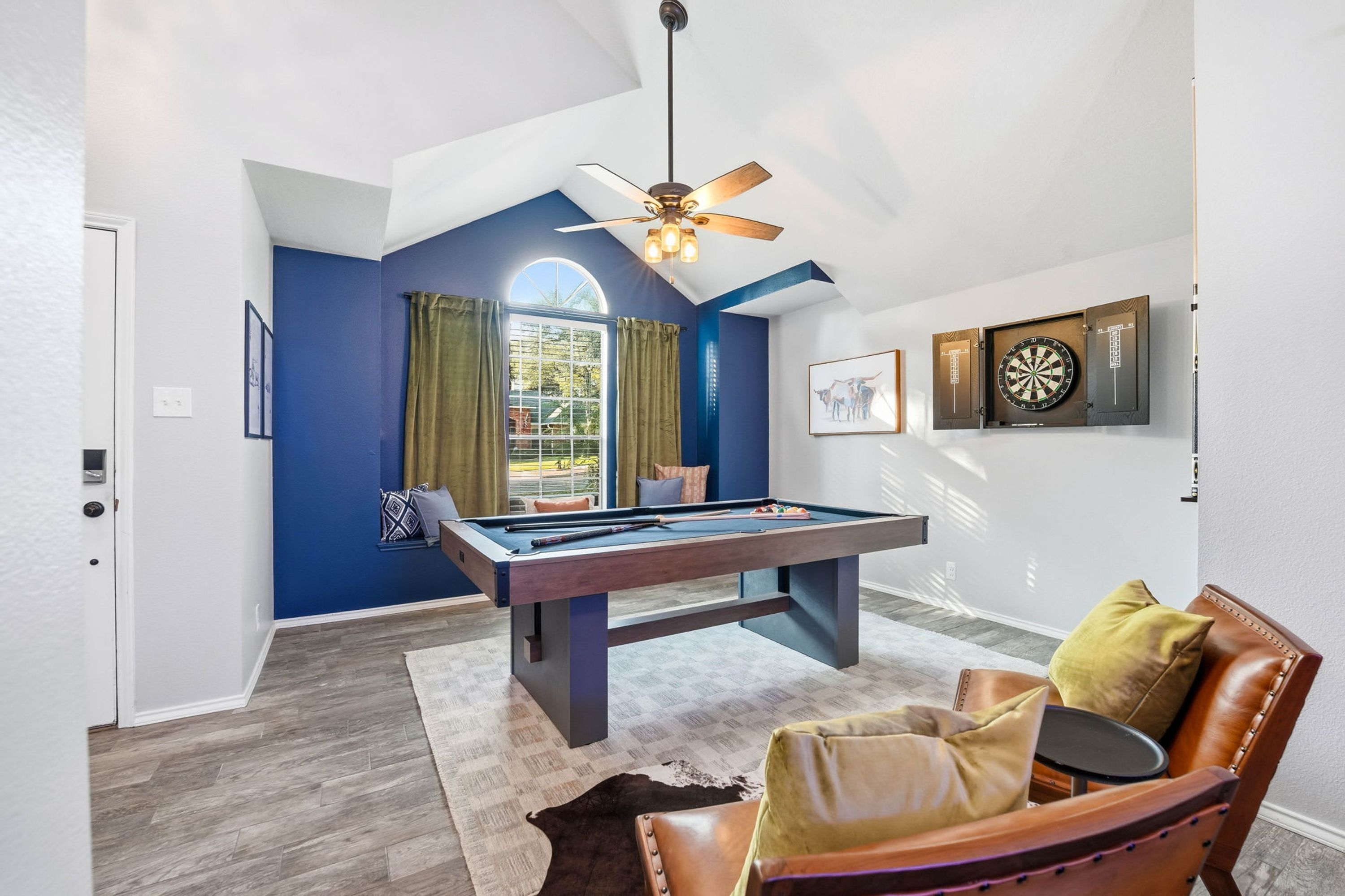 A pool table in a room with a ceiling fan, a dartboard, and a window with curtains.