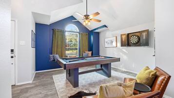 A pool table in a room with a ceiling fan, a dartboard, and a window with curtains.