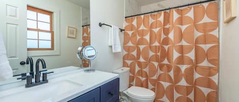 Un baño con lavamanos blanco, un espejo, inodoro y cortina de ducha con diseño geométrico.