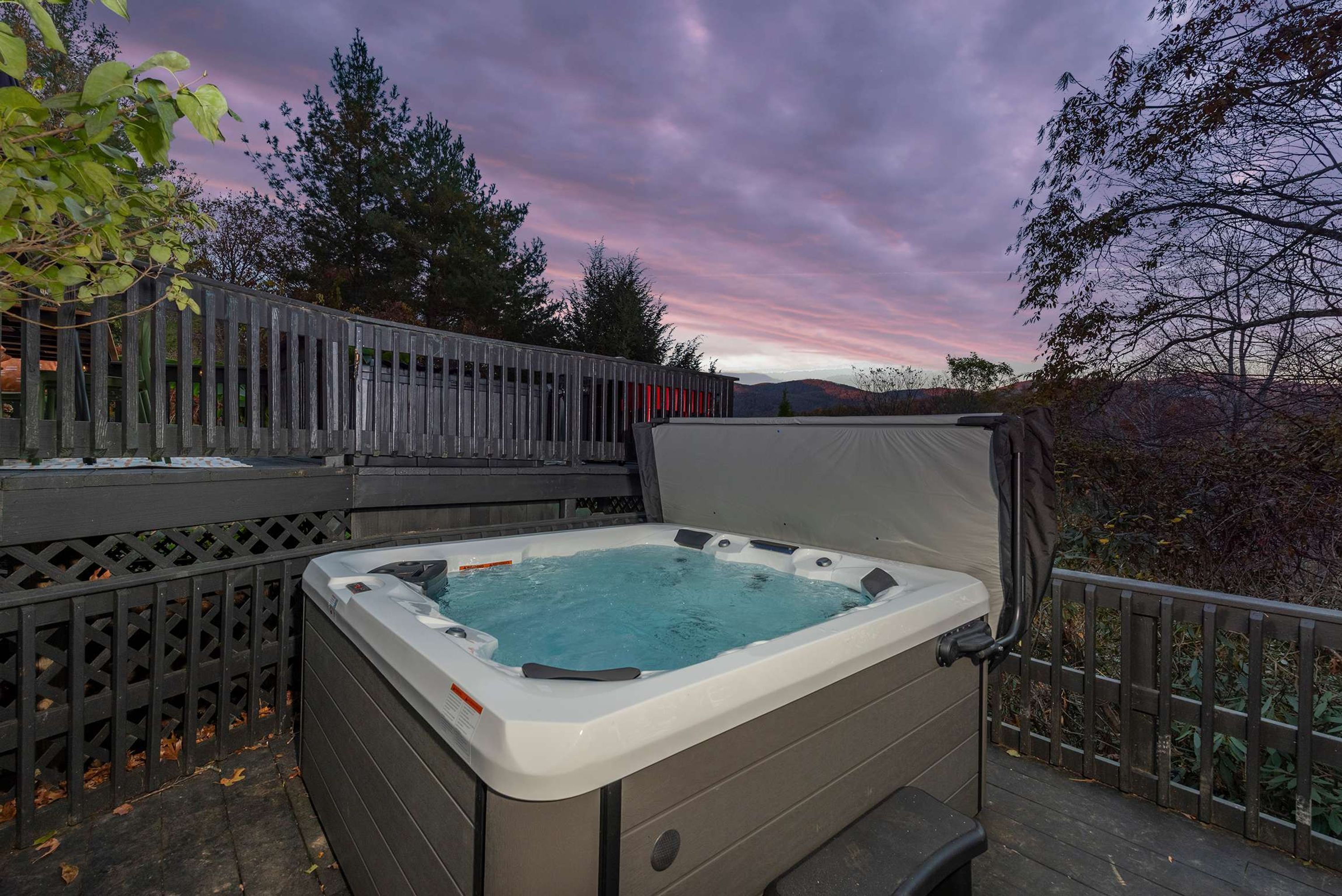 A hot tub on an outdoor deck with a view of trees and a sunset.
