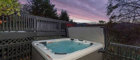 A hot tub on an outdoor deck with a view of trees and a sunset.