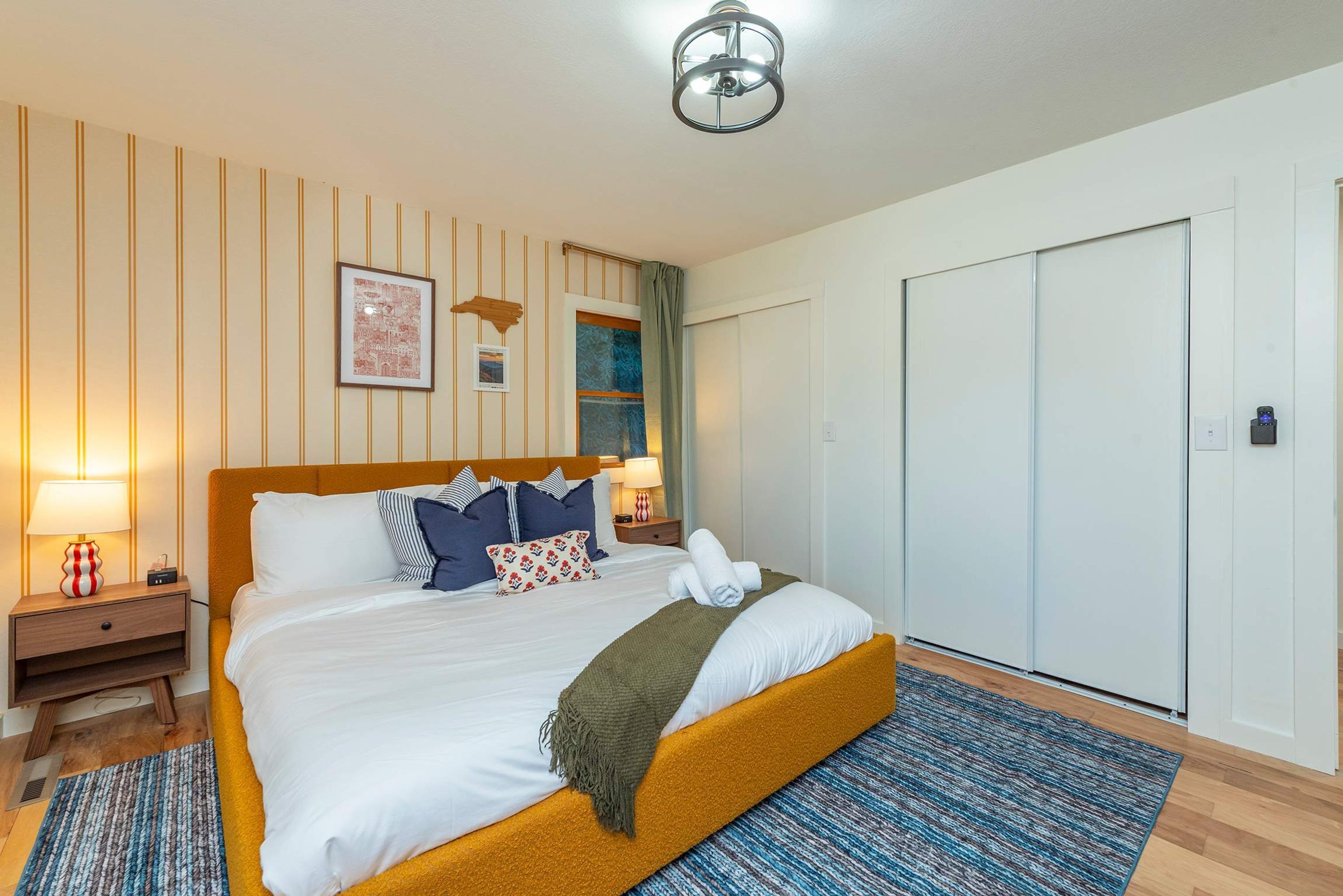 A hotel room with a bed, bedside tables, a nightstand, a lamp, a framed picture, a window with curtains, a door, and a ceiling light.