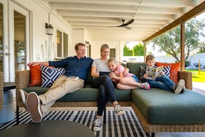 A family of four on a green sofa with a ceiling fan, a wicker table, and a view of a green lawn. - The Sunkiss Pickleball Heated Pool Hot Tub (Scottsdale)