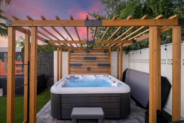 A hot tub under a wooden pergola with a brick wall and a garden in the background. - The Sunkiss Pickleball Heated Pool Hot Tub (Scottsdale)