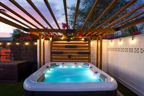 A hot tub under a wooden pergola with string lights, surrounded by a brick wall and flowering plants. - The Sunkiss Pickleball Heated Pool Hot Tub (Scottsdale)