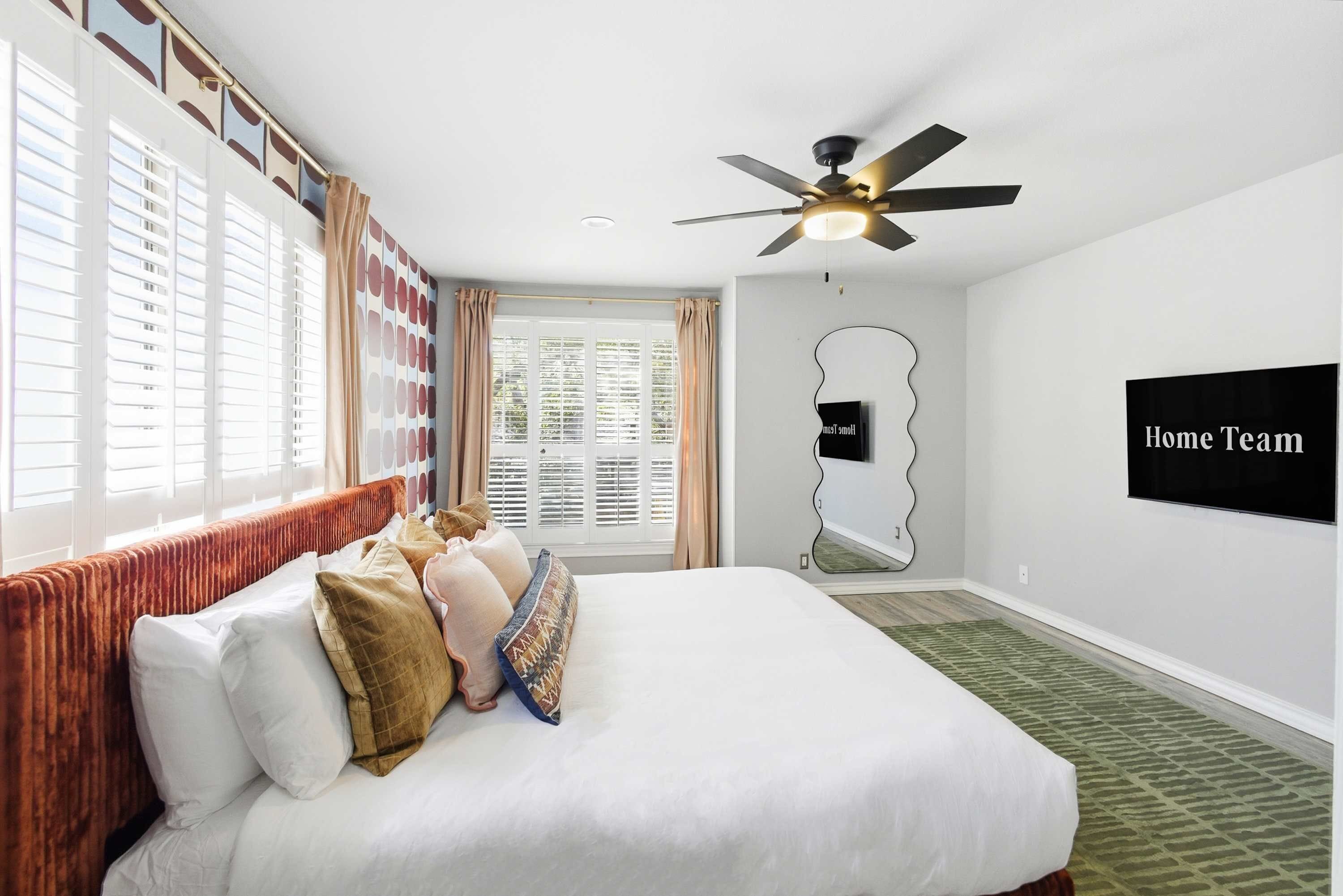 A hotel room with a large bed, a ceiling fan, a TV mounted on the wall, and a window with blinds.