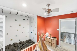 A room with a white ceiling, a ceiling fan, a red wall, a white wall with climbing holds, a wooden playpen, and a door leading to a balcony. - Ultimate Backyard Pool Game Room Kid s Room (San Antonio)