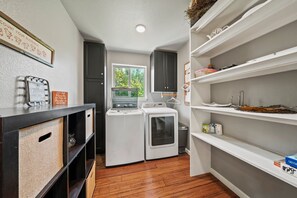A laundry room with a washing machine and dryer, a window with a view of trees, and shelves with various items. - 2 Home Getaway-pool-hot Tub-pickleball Court (Fredericksburg)