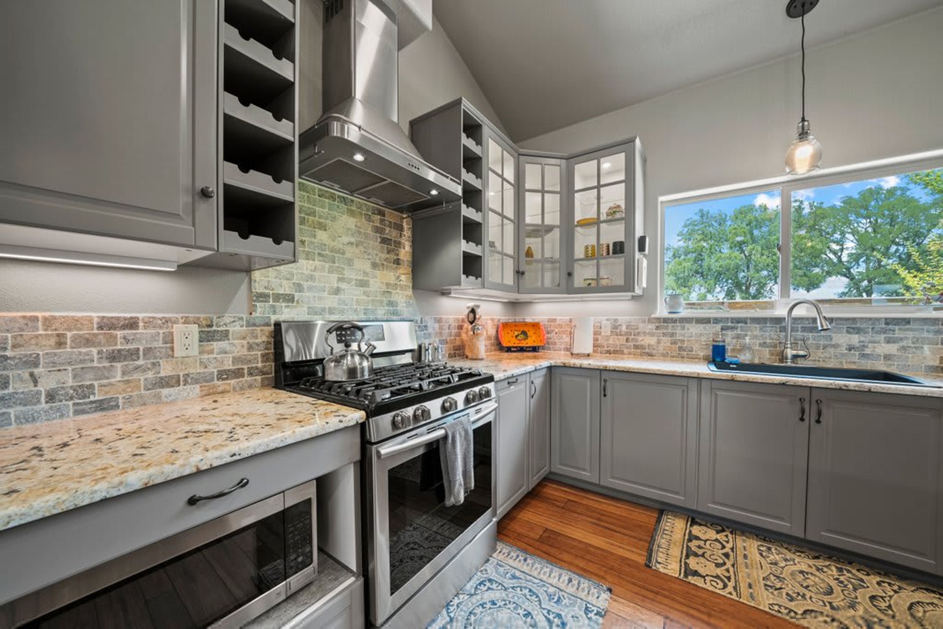A kitchen with a gas stove, oven, and wine rack, featuring a stone backsplash and grey cabinetry.