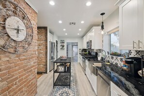 A modern kitchen with a brick accent wall, a large clock, and a central island. - The Glow-up House Pool Spa Glam Room (Phoenix)