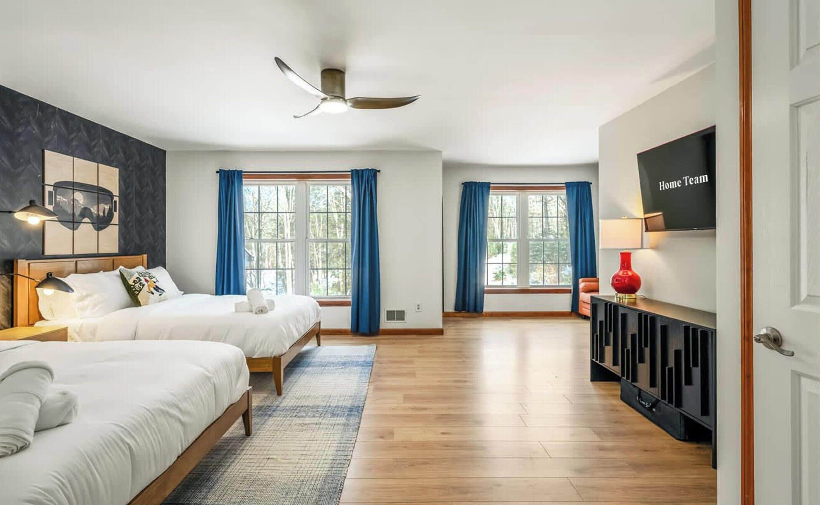 A hotel room with two beds, a ceiling fan, large windows with blue curtains, a flat-screen TV, and a red vase on a console table.