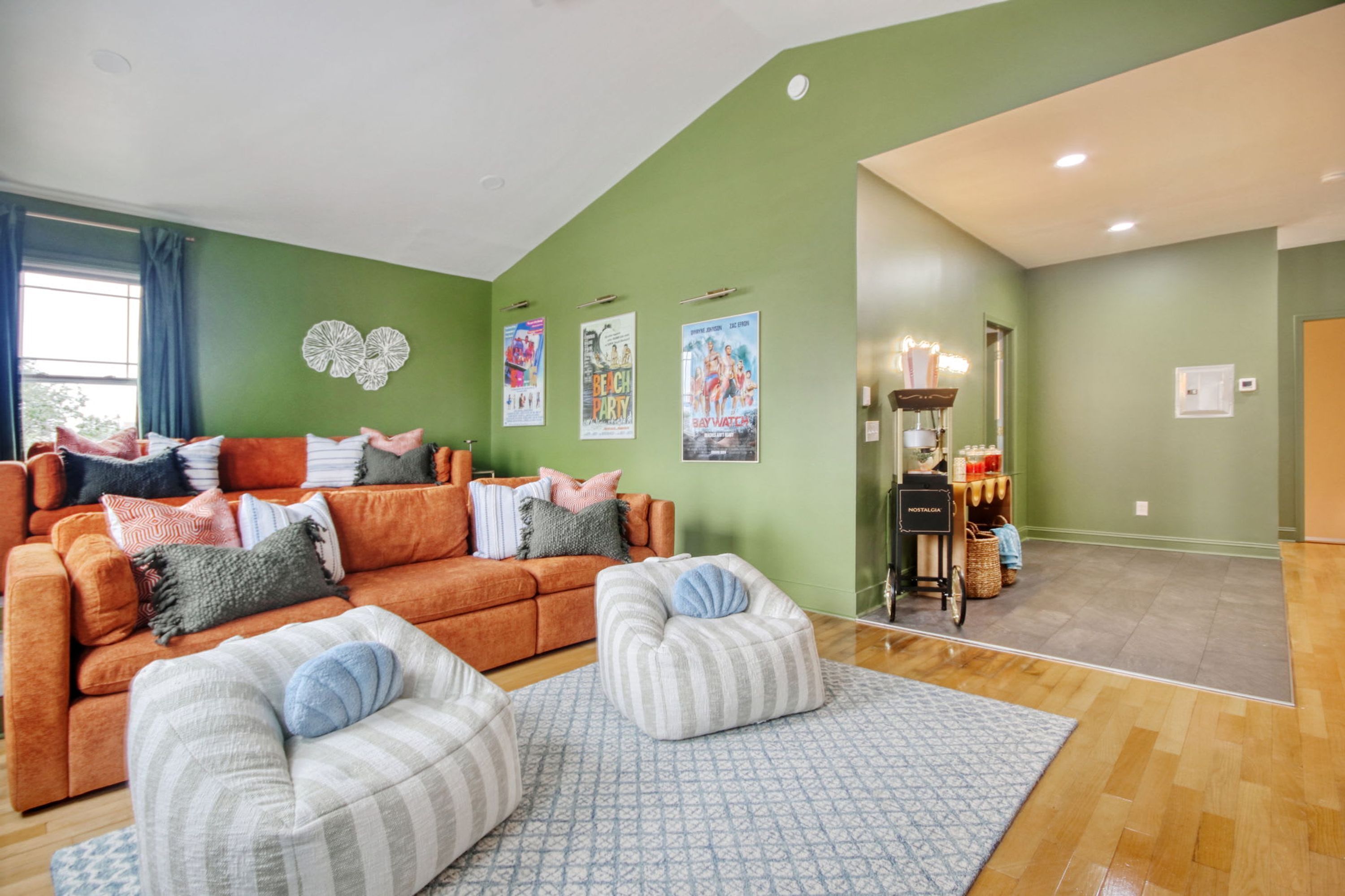 Una sala de estar con pared verde, un sofá, dos otomanas y una mesa de centro. Hay carteles en la pared y un mueble bajo con objetos encima.