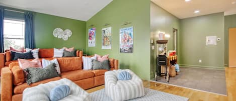 A living room with a green wall, a sofa, two ottomans, and a coffee table. There are posters on the wall and a small cabinet with items on it.