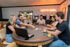 A group of people playing cards around a table in a room with a neon sign reading 'Surf & Game Night Games'. - Golf Sim Theater Games Walk to Beach Sleeps 28 (North Myrtle Beach)