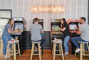 Four people playing arcade games in a room with a neon sign that reads 'Sandy Toads & Game Night Games'. - Golf Sim Theater Games Walk to Beach Sleeps 28 (North Myrtle Beach)