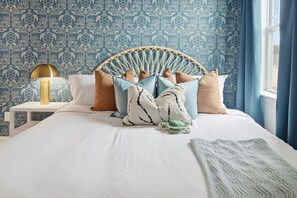 A bedroom with a patterned wallpaper, a rattan headboard, multiple pillows, a bedside lamp, and a window with curtains. - Golf Sim Theater Games Walk to Beach Sleeps 28 (North Myrtle Beach)