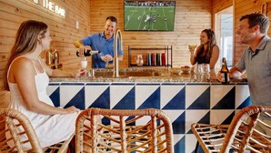 Zona de bar amb una pantalla de televisió que mostra un partit de futbol, tamborets de bar i una barra on un barman prepara una beguda.