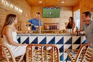 A bar area with a television screen displaying a football game, bar stools, and a counter where a bartender is preparing a drink. - Golf Sim Theater Games Walk to Beach Sleeps 28 (North Myrtle Beach)