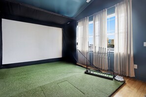 A room with a large white projection screen, a green artificial turf floor, a metal staircase with a mesh railing, and a window with sheer curtains. - Golf Sim Theater Games Walk to Beach Sleeps 28 (North Myrtle Beach)