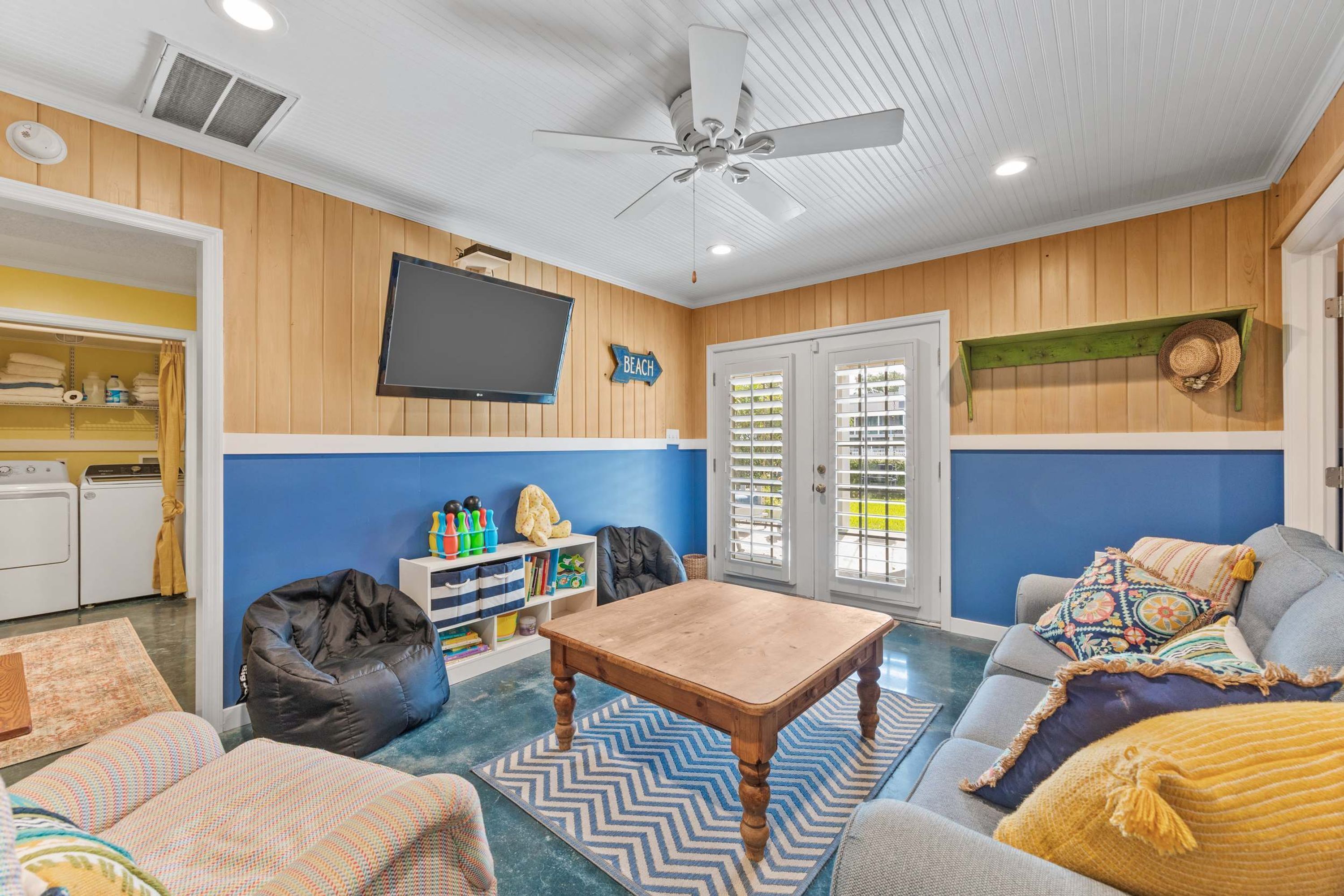 A room with a ceiling fan, a television mounted on the wall, a door with a view to the outside, and a seating area with a table and chairs.