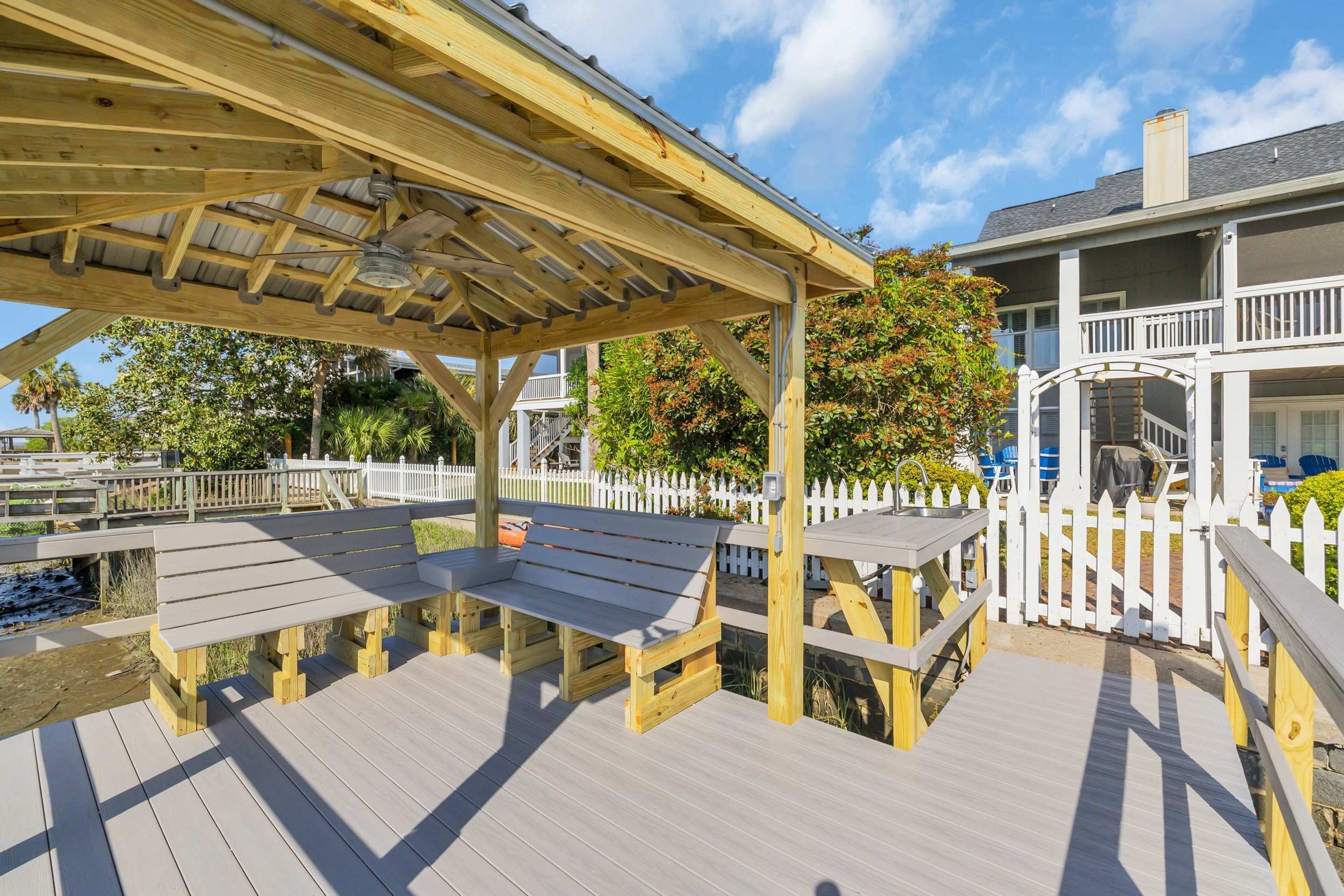 A wooden deck with benches and a pergola, surrounded by a white picket fence and a house with a balcony.