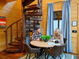 Un interior de madera con una escalera de caracol, una mesa redonda con sillas y un ventanal con cortinas.