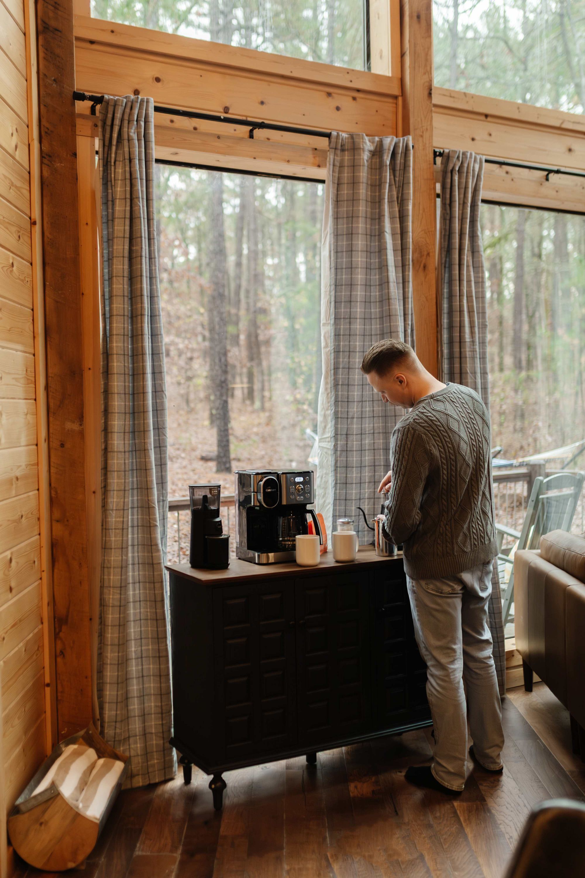 Una persona de pie junto a una ventana con cortinas, una cafetera y una taza sobre un aparador de madera.