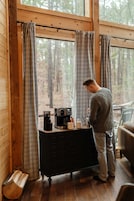 Una persona de pie junto a una ventana con cortinas, una cafetera y una taza sobre un aparador de madera.