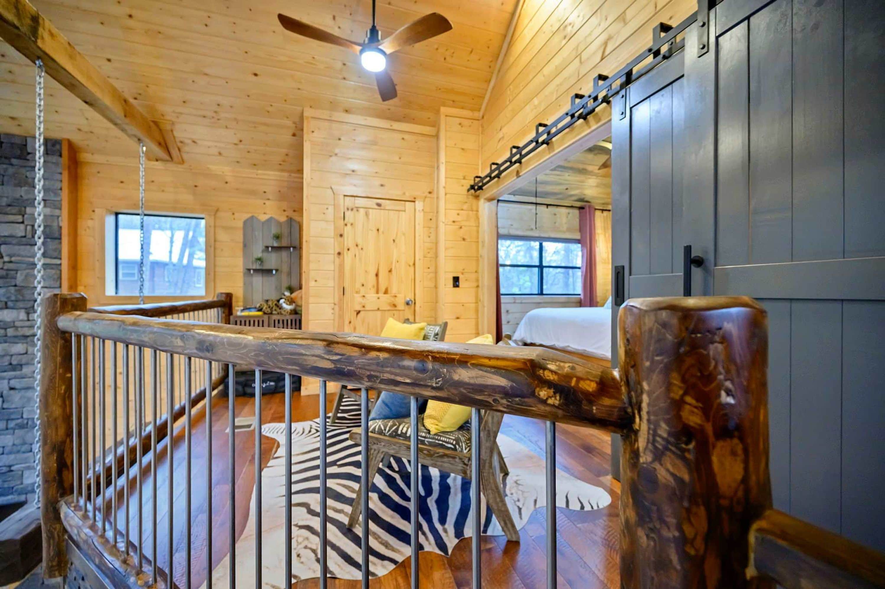 Interior de una cabaña de madera con balcón, ventilador de techo y una cama con alfombra de estampado de cebra.