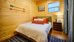 A cozy wooden cabin bedroom with a bed, pillows, a nightstand, a window, and a painting on the wall. - Retro Retreat Stargazing Dome Sauna Hot Tub (Broken Bow)