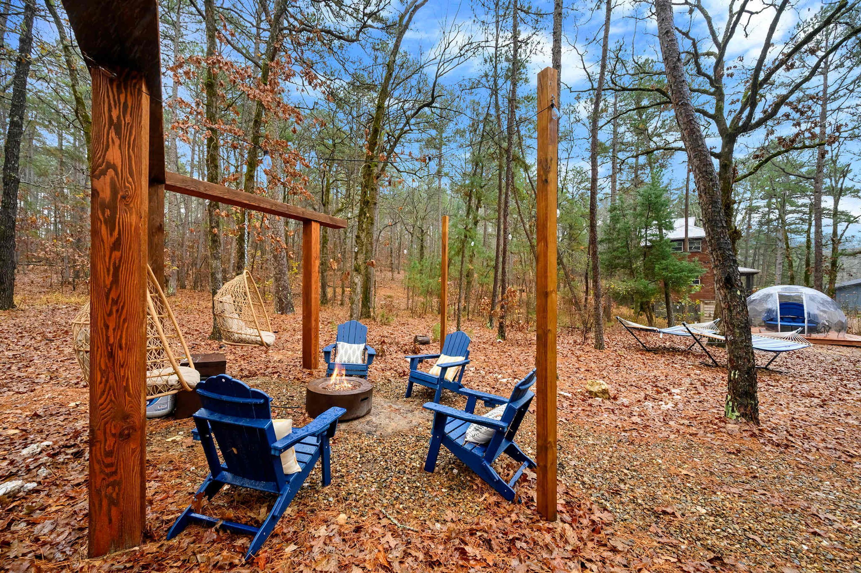 Un campamento rodeado de bosque con un pozo para fuego, sillas Adirondack y una tienda de campaña.