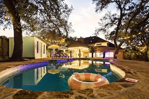 A swimming pool with a lounge area and umbrellas, surrounded by trees and a house. - Gathering House Movie Game Room Hot Tub Pool (San Antonio)