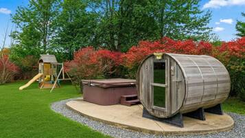 A wooden hot tub on a circular platform, a wooden playhouse with a slide, and a garden with red foliage.