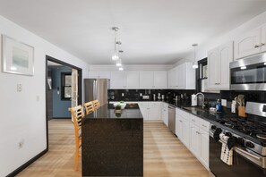 A modern kitchen with white cabinets, a black countertop, stainless steel appliances, and a dining area with a wooden table and chairs. - Sleeps 14 Hot Tub Sauna Game Room Fire Pit (Long Pond)