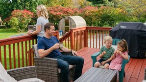 Eine Familie auf einer Holzterrasse mit einem Grill und einem kleinen Schuppen im Hintergrund.