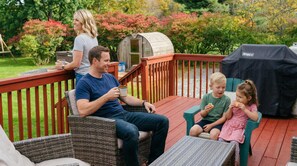 Eine Familie auf einer Holzterrasse mit einem Grill und einem kleinen Schuppen im Hintergrund.