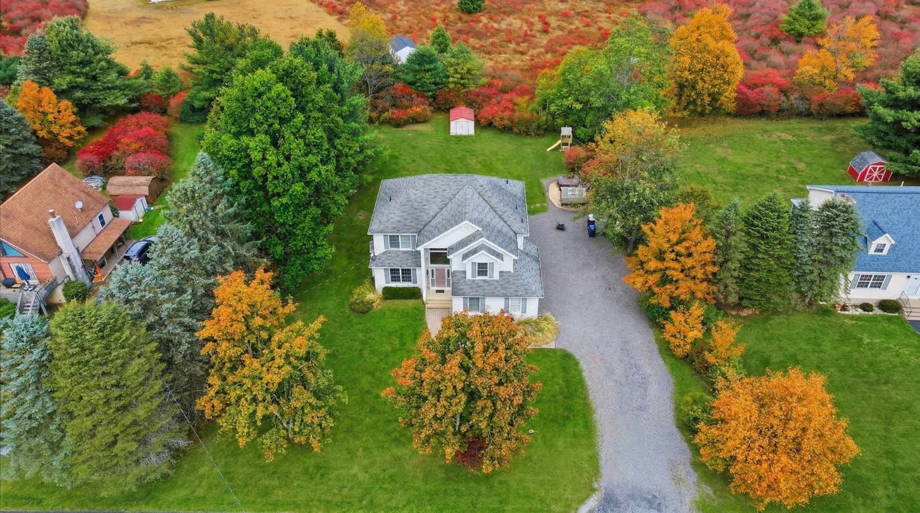 A residential area with a two-story house, a driveway, and surrounding trees with autumn foliage.