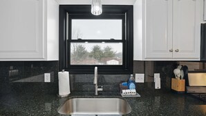 Eine Küche mit Spülbecken aus Edelstahl, einem Fenster mit Blick auf Bäume und ein Gebäude sowie einer Arbeitsplatte mit einer Flasche Geschirrspülmittel und einem Handtuch.