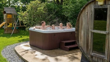 Un jacuzzi con revestimiento de madera y escalones de acceso, rodeado de un césped y una casita de juegos de madera.