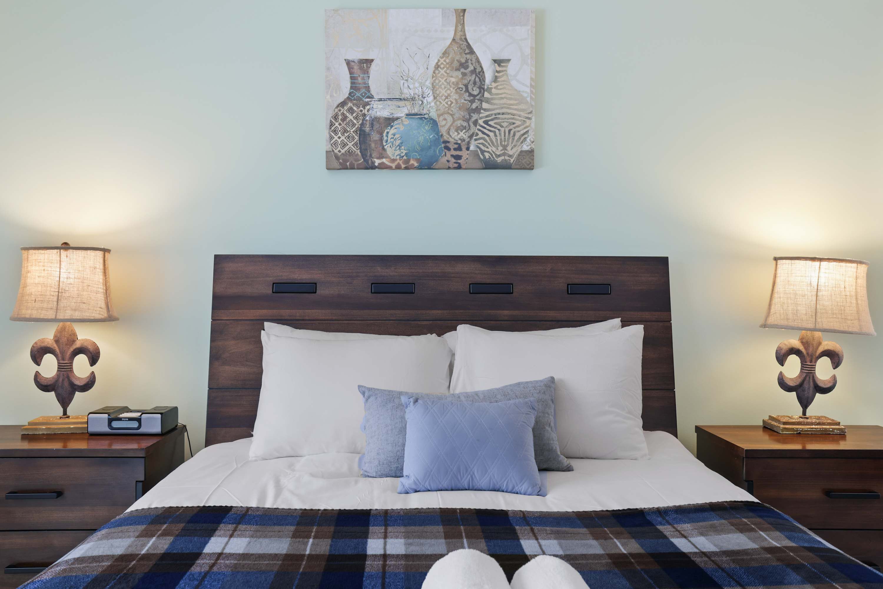 A neatly made bed with white and blue bedding, flanked by two bedside tables with lamps, and a painting above.