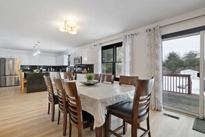 A modern kitchen with white cabinets, a dining table with chairs, and a view of the outdoors through a large window. - Sleeps 14 Hot Tub Sauna Game Room Fire Pit (Long Pond)