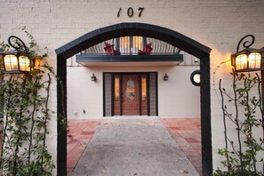 Ein Gebäudeeingang mit der Nummer 107 darüber, eine Holztür mit Glasfeldern, ein Balkon mit roten Stühlen und zwei an der Wand befestigte Laternen.