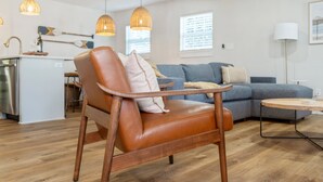 Un salon moderne avec un fauteuil en cuir, un canapé bleu, un plancher en bois, des luminaires suspendus et un coin cuisine à l’arrière-plan.
