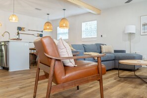 Ein modernes Wohnzimmer mit Ledersessel, blauem Sofa, Holzböden, Pendelleuchten und einer Küchenzeile im Hintergrund.