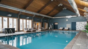 An indoor swimming pool with clear blue water, wooden ceiling beams, and a view of the outside through large windows. - Luxury Lakeview Estate w Indoor Pool Golf Sim (Ithaca)