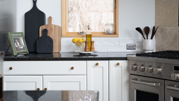 A kitchen with white cabinets, a black countertop, a stove, and a window with a view of trees. - Luxury Lakeview Estate w Indoor Pool Golf Sim (Ithaca)