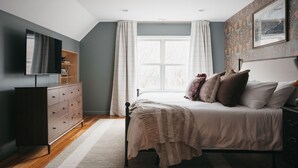 A bedroom with a bed, a dresser, a television, and a window with curtains. - Luxury Lakeview Estate w Indoor Pool Golf Sim (Ithaca)