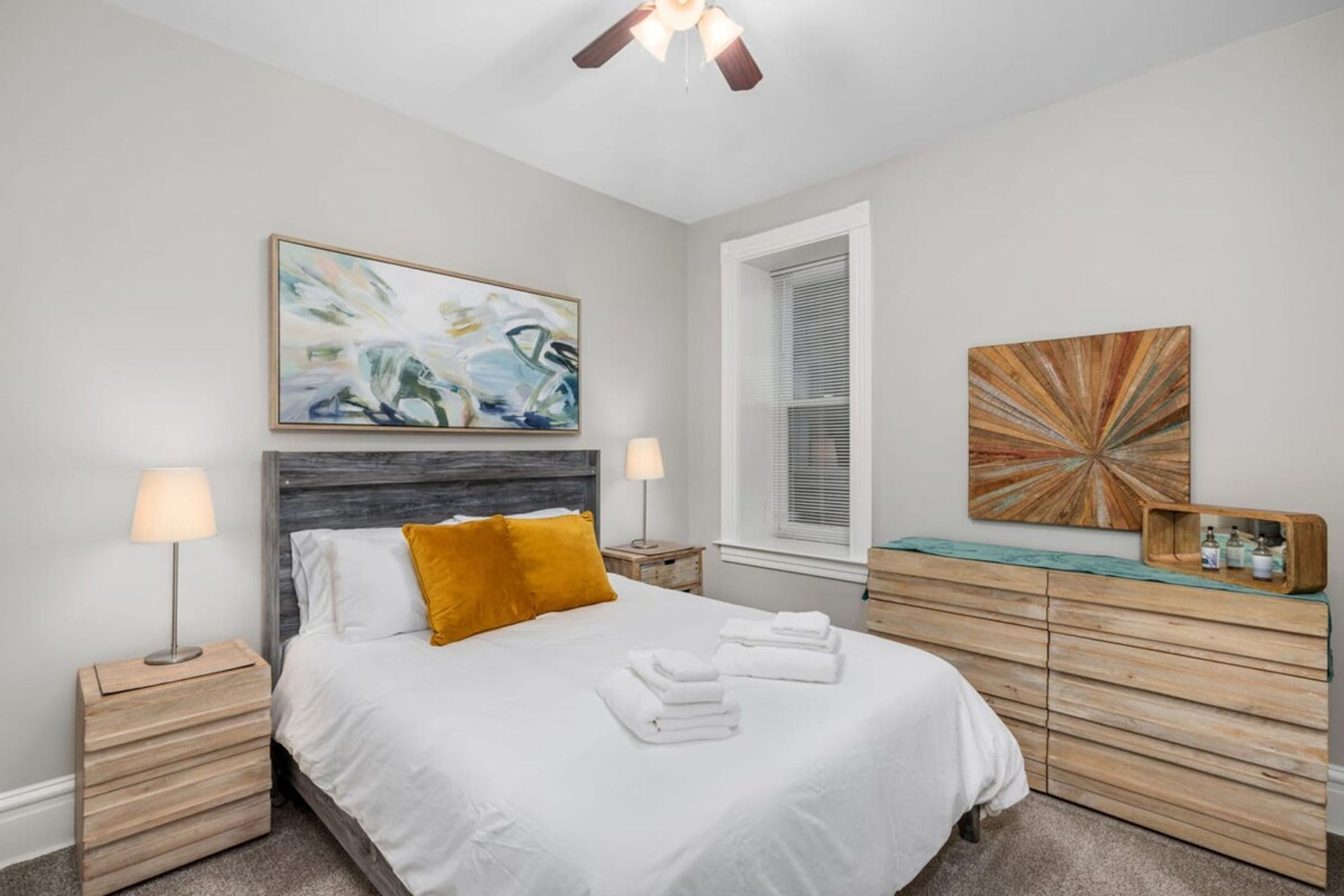Une chambre à coucher comprenant un lit, deux tables de chevet, une commode, une coiffeuse, une fenêtre avec des stores et deux lampes fixées au mur.