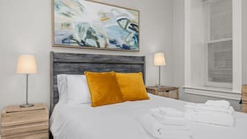 A neatly made bed with white bedding and a mustard-colored pillow, flanked by two bedside tables with lamps, in a room with a large abstract painting above the bed and a window with blinds to the right.